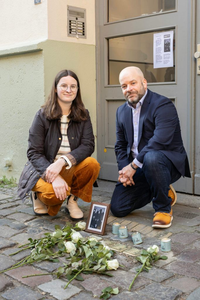 A woman and man kneeling at a memorial with a framed photo, candles and white roses on it.