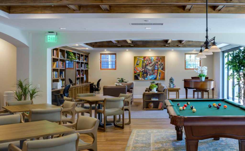 A room with several tables and chairs. Bookcases, couches and a painting are in the back of the room. A pool table is on the right side of the room.