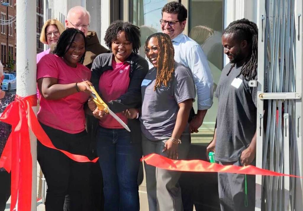 Several men and women laughing and smiling. One woman is holding a giant pair of scissors and has just cut a ribbon.