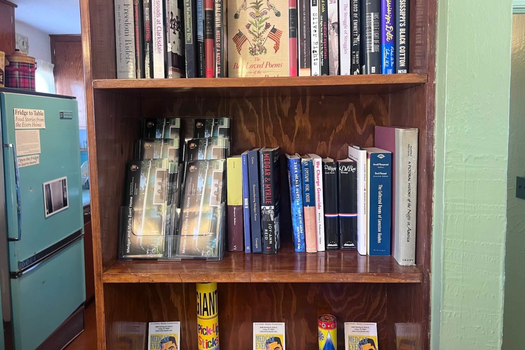 brochures with a photo of the Evers home and a photo of Medgar and Myrlie Evers on its cover sit on a bookshelf next to several books on Black history