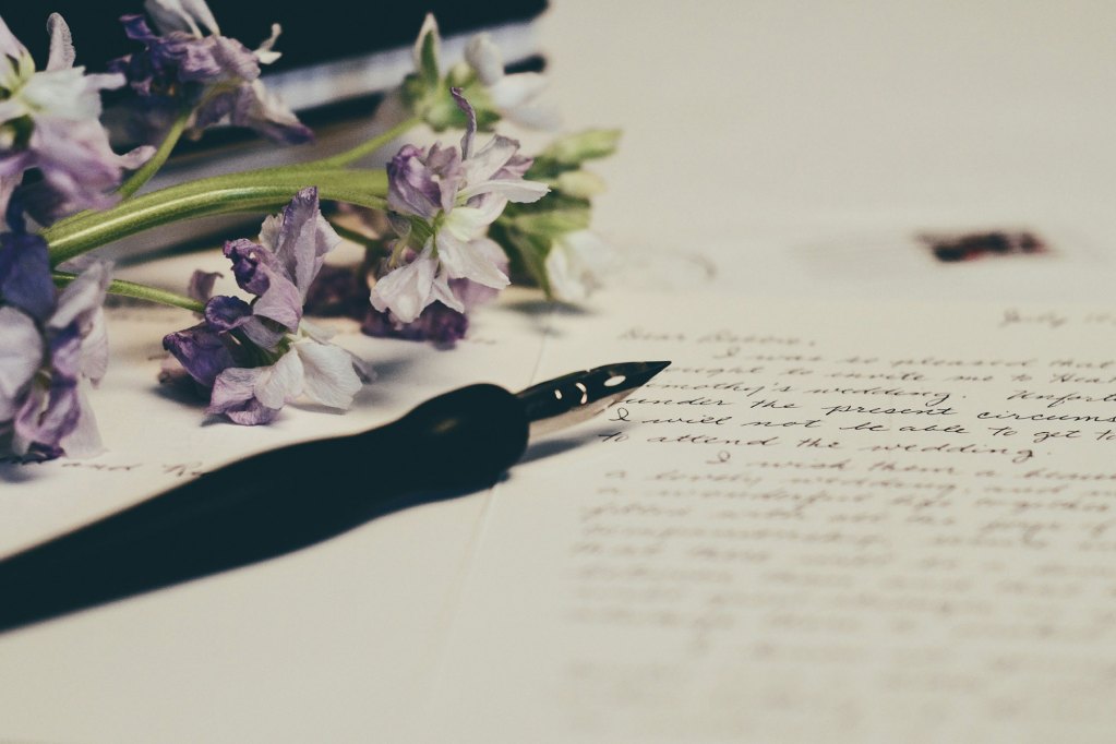 A black fountain pen next to a set of purple flowers. The pen is laid on a sheet of paper with writing on it.