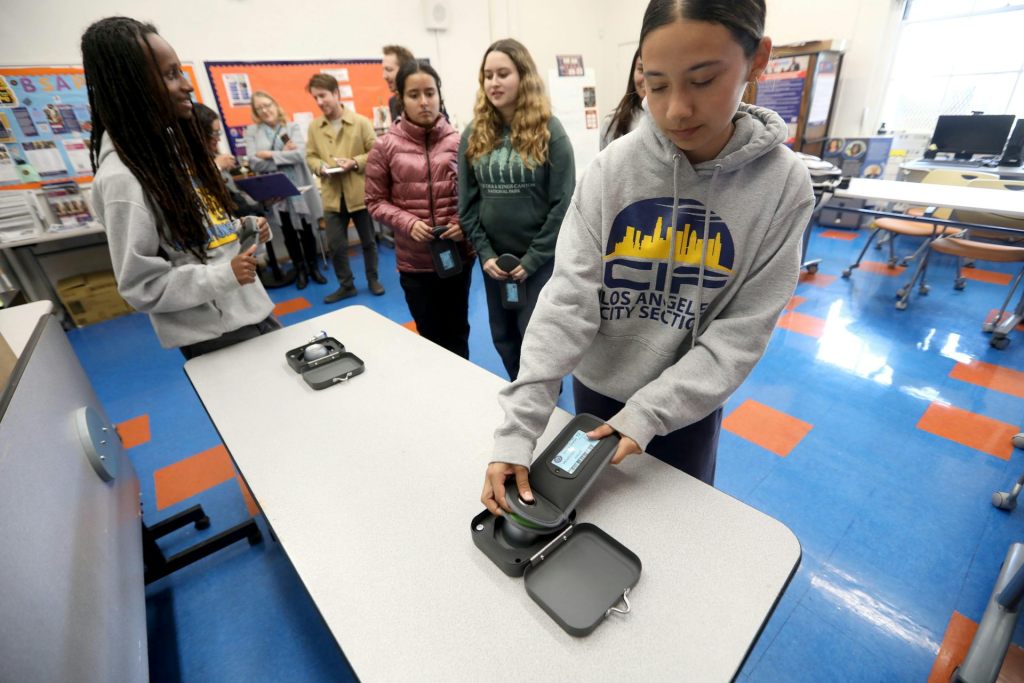 A female student places her cell phone into a locked case on a table.