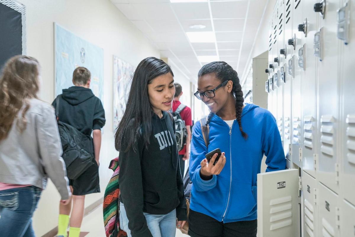 Two female students looking at a cell phone screen while standing in a school hallway.