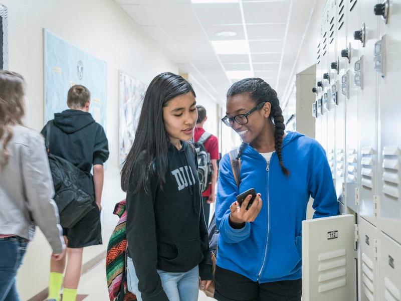 Two female students looking at a cell phone screen while standing in a school hallway.
