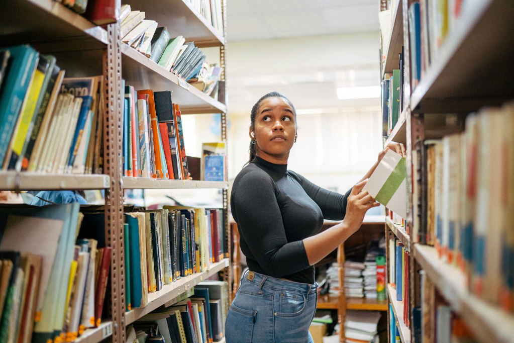 A woman standing in between shelves of books in a library, picking books and looking off into the distance above