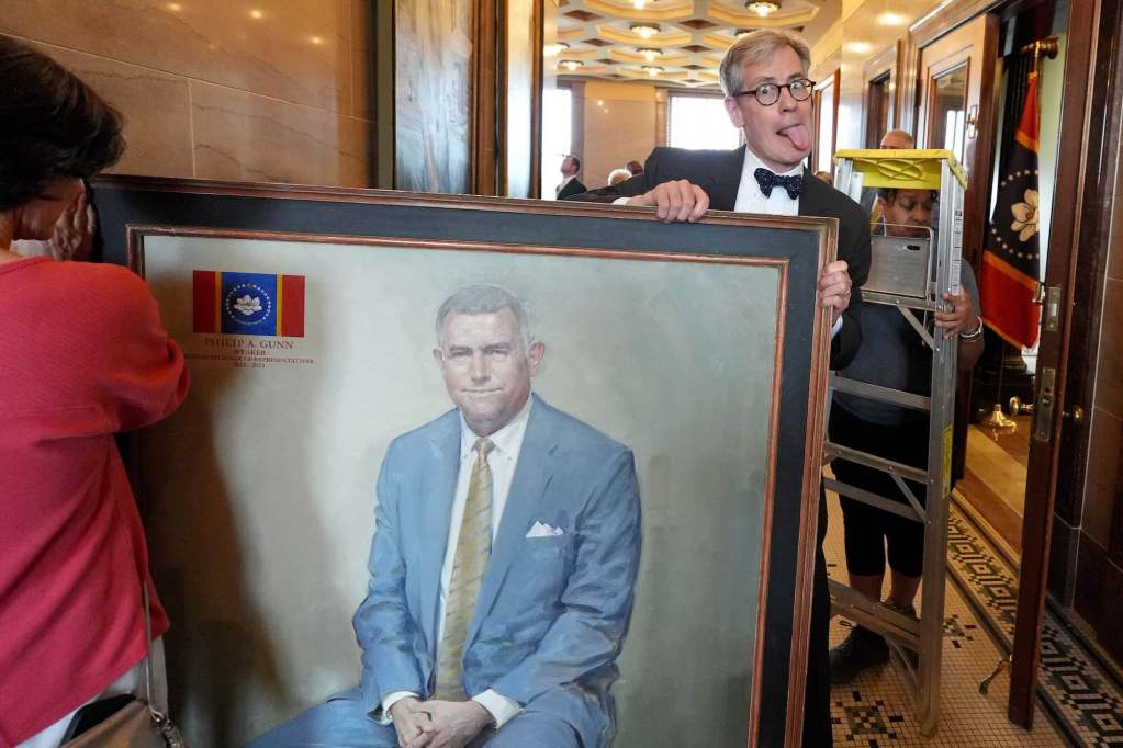 A man holds a portrait from one end and makes a silly face as someone holds the other end