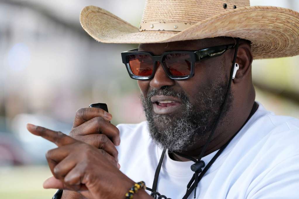 A man wearing a straw cowboy hat and sunglasses speaks outside