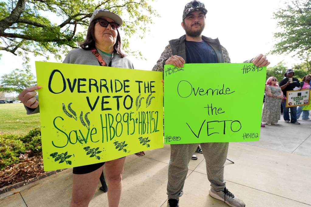 Two people hold signs outside. A yellow sign reads 'Override the Veto, Save HB895 & HB1152' while the green sign beside it reads 'Override the Veto'