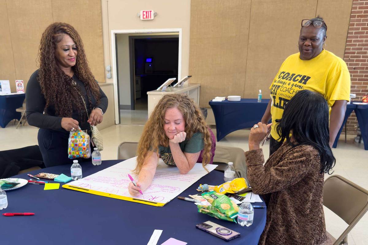 Four women surrounding a table, with one woman seated and writing on a large easel pad.
