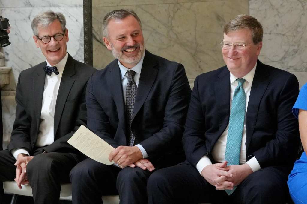 Three men, including Tate Reeves, sit beside each other smiling