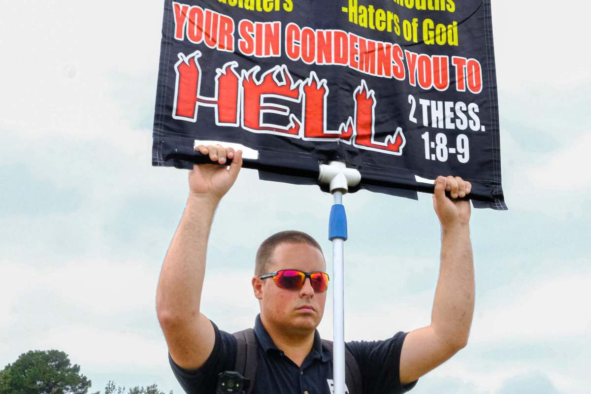 A man stands outside and holds up a black sign on a pole. Some text that's seen reads 'Your sin condemns you to hell'