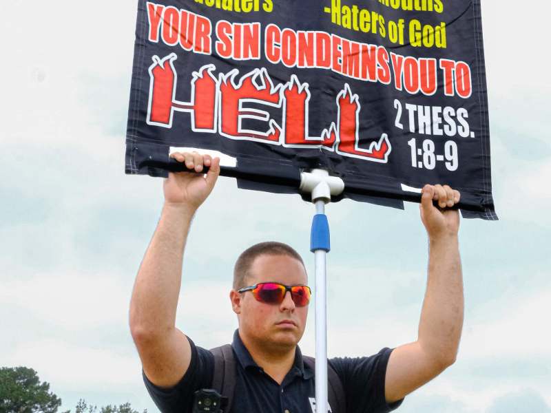 A man stands outside and holds up a black sign on a pole. Some text that's seen reads 'Your sin condemns you to hell'