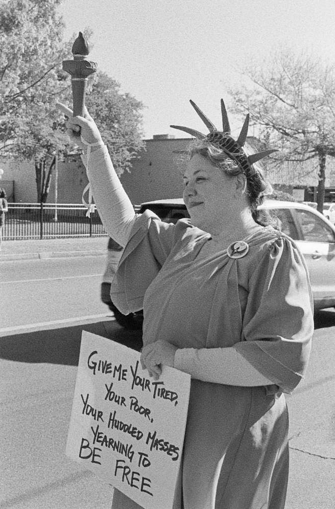 A woman dressed as the statue of liberty with a sign that reads 'Give me your tired, your poor, your huddled masses yearning to be free'