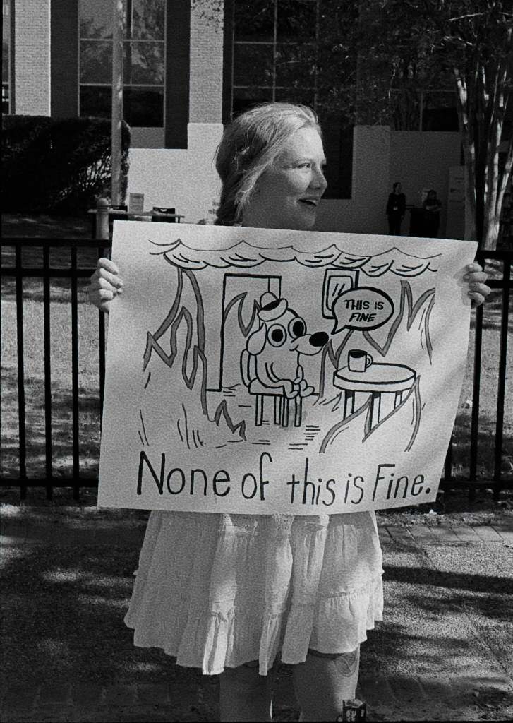 A protester holds up a sign that reads 'None of this is Fine' beneath art of a dog man sitting in a room that's on fire