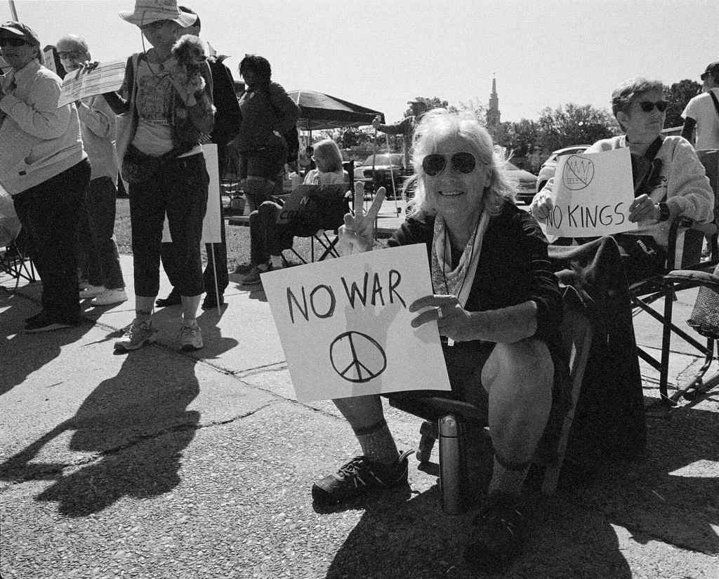 A seated woman in a protest crowd carries a simple sign that reads 'No War' with a peace symbol