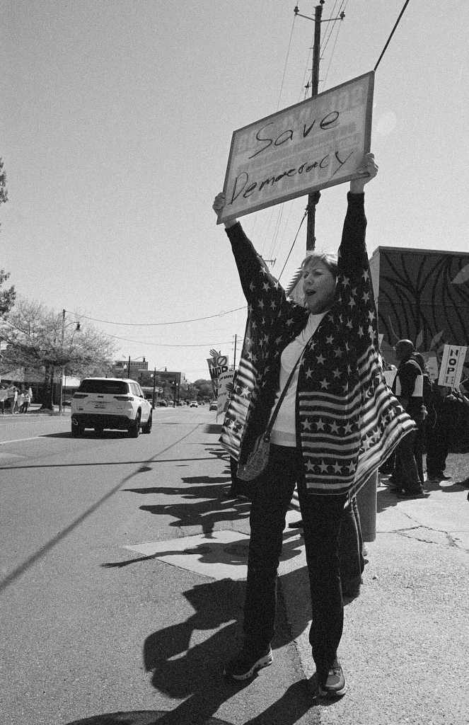 A person on the side of the road holds a sign that reads 'SAVE DEMOCRACY'