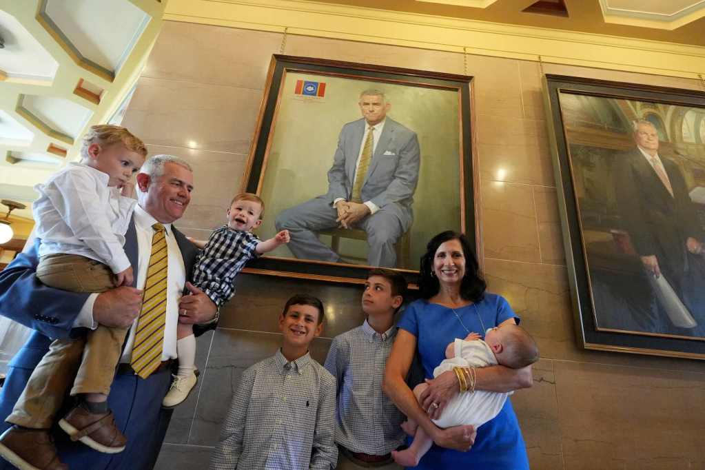 Philip Gunn, holding two small children, smiles and poses beside his portrait with other family members