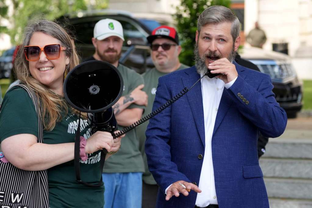 A bearded man in a blue suit speaks into a megaphone at an outside event