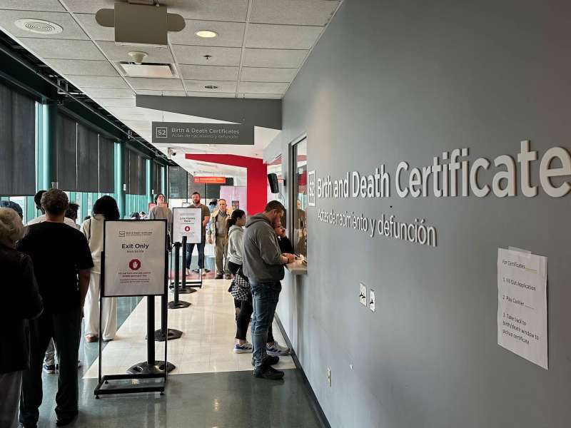 Several people in a line inside a building, with one person standing at a service window filling out paperwork.