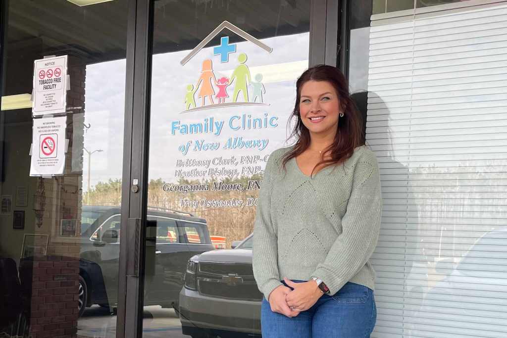 A woman with brown hair stands outside of a door labeled 'Family Clinic of New Albany'