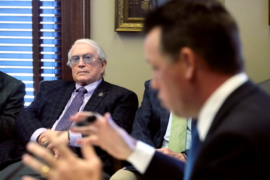 A man with white hair and glasses sits and listens to another man speaking.