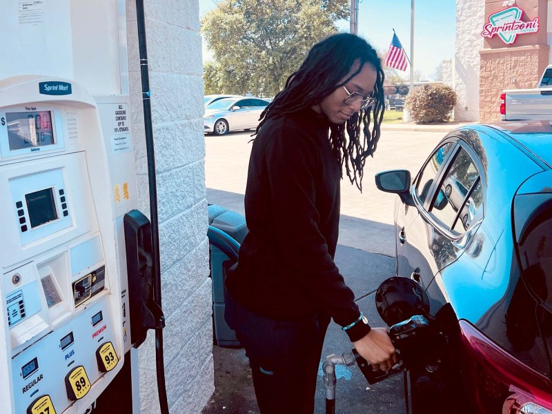 A black woman with locs pumps gas into a black car