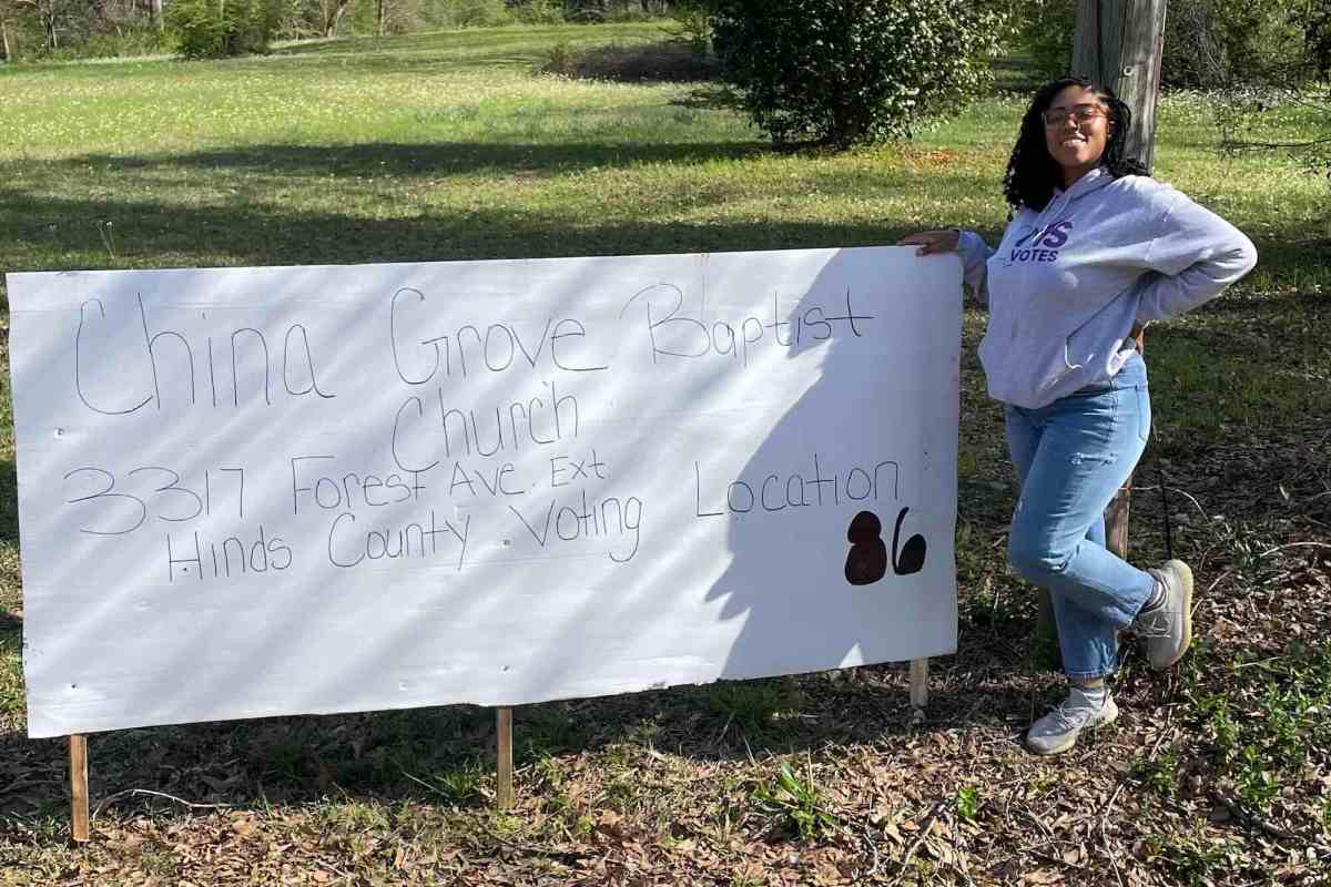 A woman stands next to a hand written sign for the China Grove Baptist Church Hinds County Voting Location 86