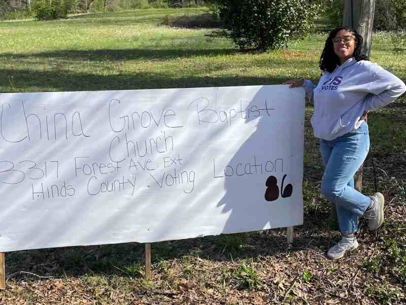 A woman stands next to a hand written sign for the China Grove Baptist Church Hinds County Voting Location 86