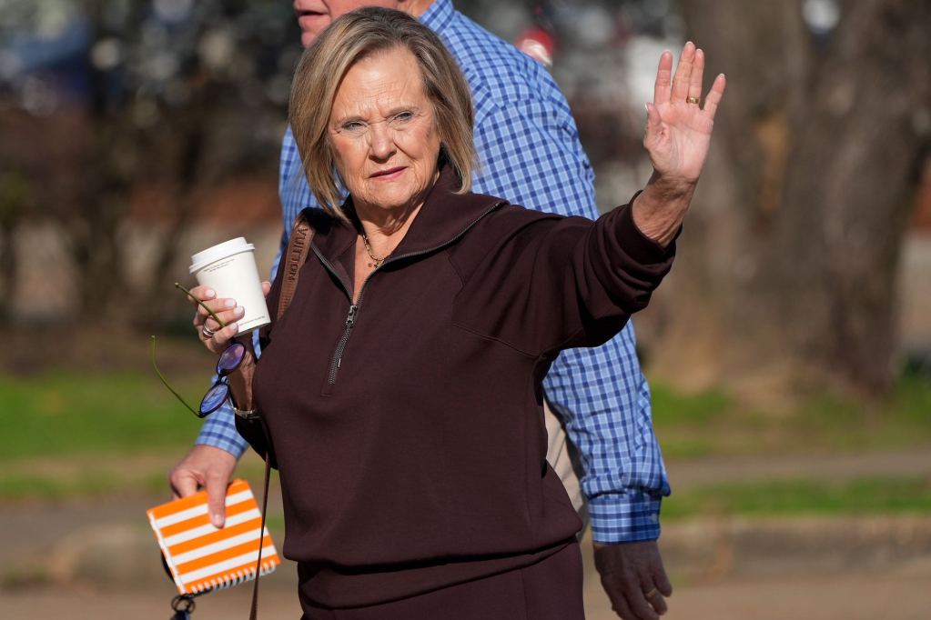 A woman holding her glasses and a coffee cup waves to someone out of view