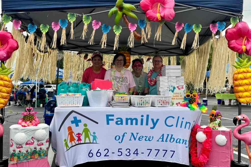 Four people stand behind a sign for 'Family Clinic of New Albany' under a tent with tropical decorations