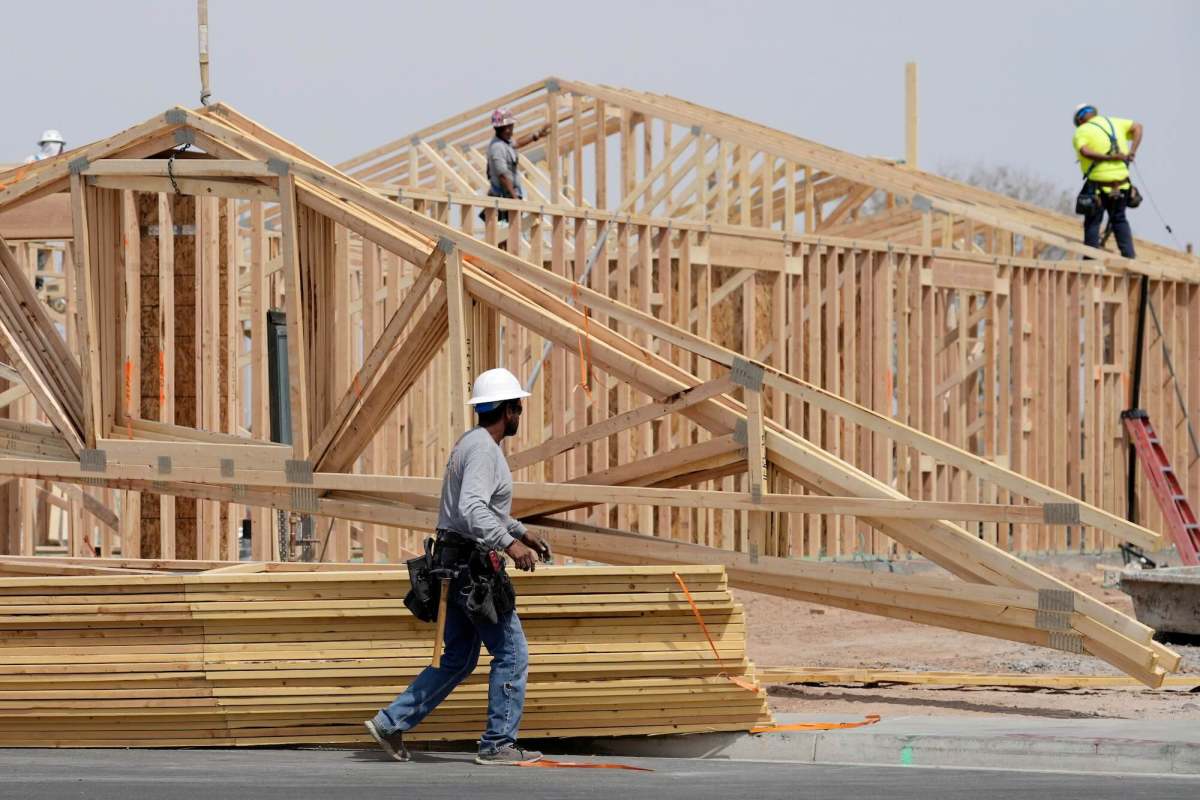 A construction worker wearing a hard hat walking in front of stacks of wooden planks and a house under construction. Two other workers are in the background.
