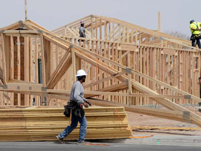 A construction worker wearing a hard hat walking in front of stacks of wooden planks and a house under construction. Two other workers are in the background.