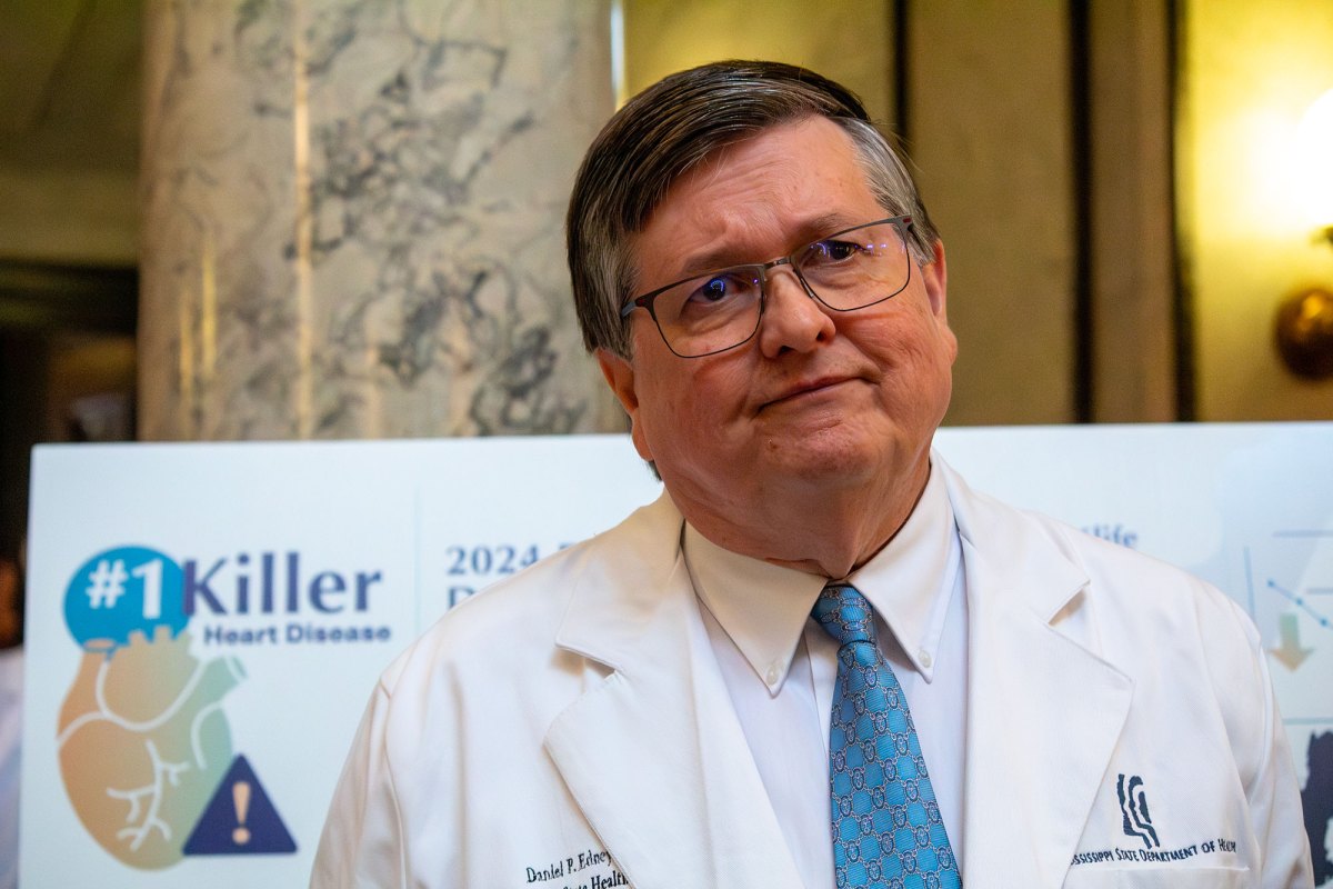 a man in glasses and da white coat that says "Daniel P Edney" on one side and "Mississippi State Department of Health" on the other stands inside a building with marble support columns listening