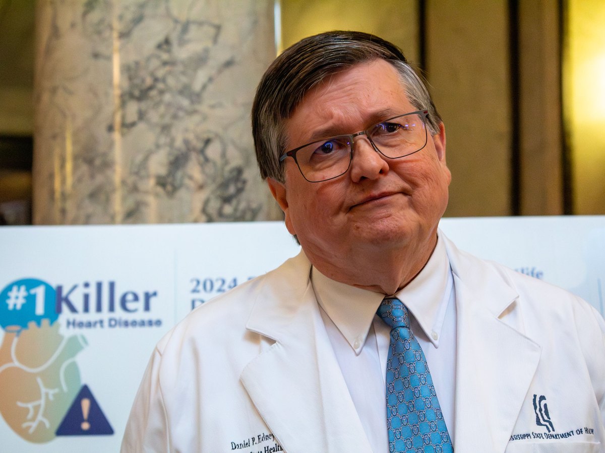 a man in glasses and da white coat that says "Daniel P Edney" on one side and "Mississippi State Department of Health" on the other stands inside a building with marble support columns listening