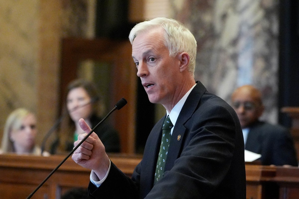 David Blount in a dark suit speaks, hand gesturing, in front of the senate chamber