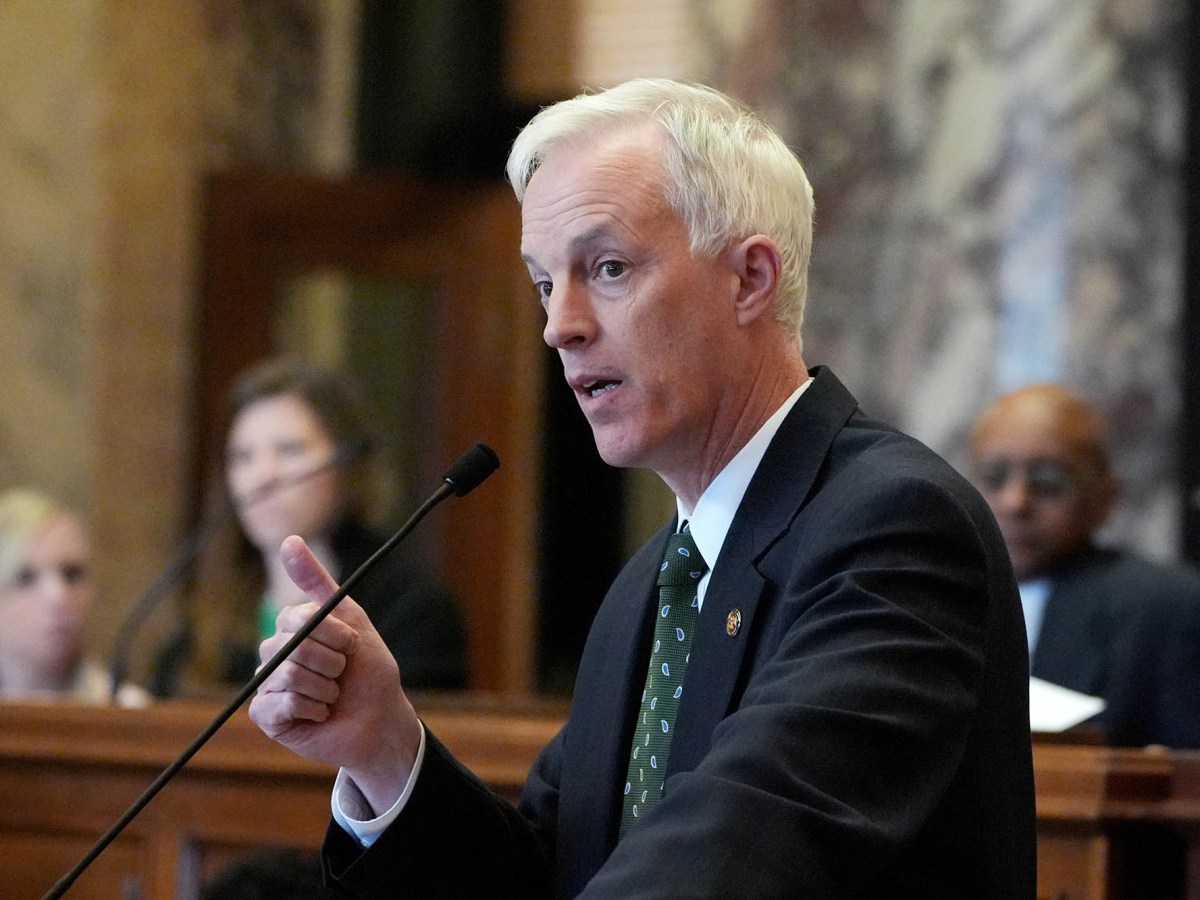 David Blount in a dark suit speaks, hand gesturing, in front of the senate chamber