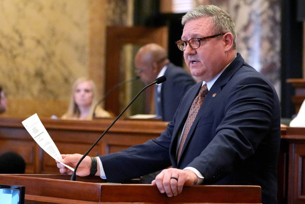Wearing reading glasses, Dennis DeBar stands at a podium and speaks while holding a piece of paper in his hand.