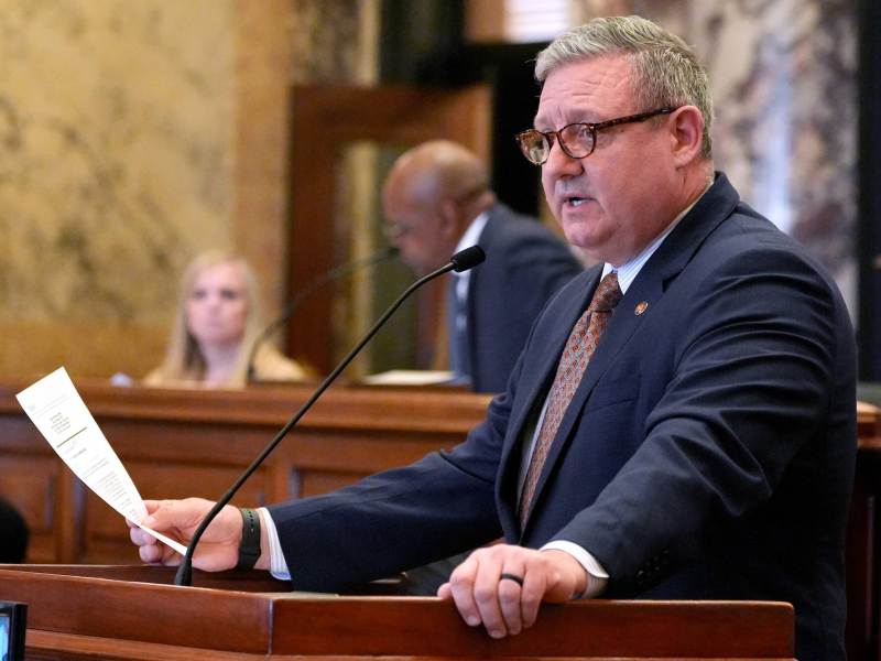 Wearing reading glasses, Dennis DeBar stands at a podium and speaks while holding a piece of paper in his hand.