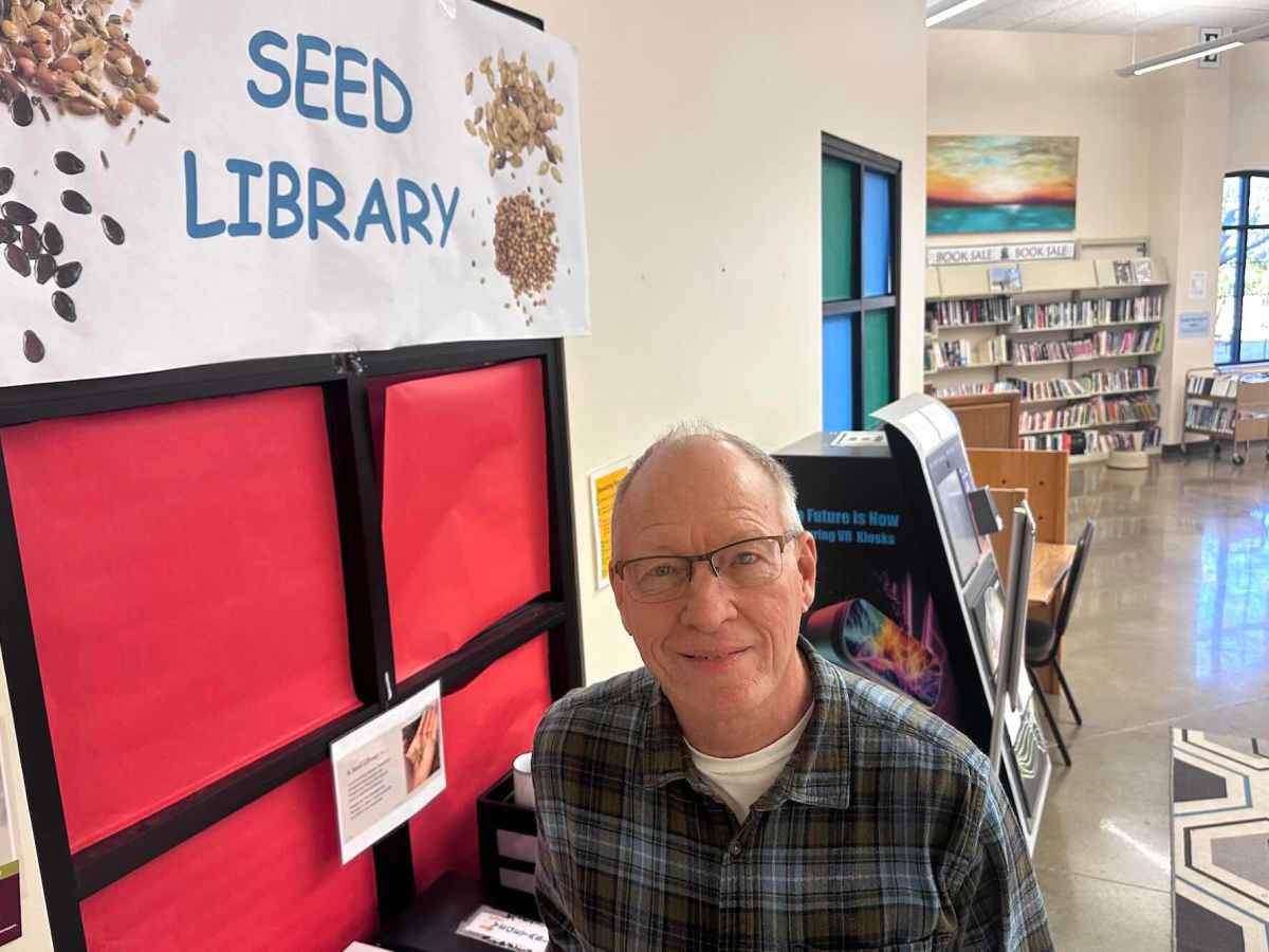A man in a green plaid shirt and glasses looks up at hte camera. A sign for 'Seed Library' is visible to the left, and library shelves to the right