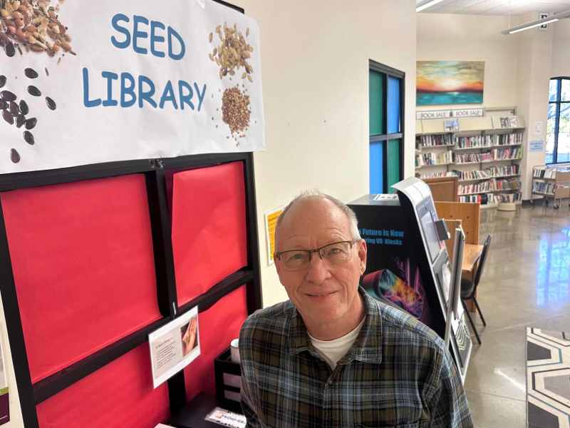 A man in a green plaid shirt and glasses looks up at hte camera. A sign for 'Seed Library' is visible to the left, and library shelves to the right