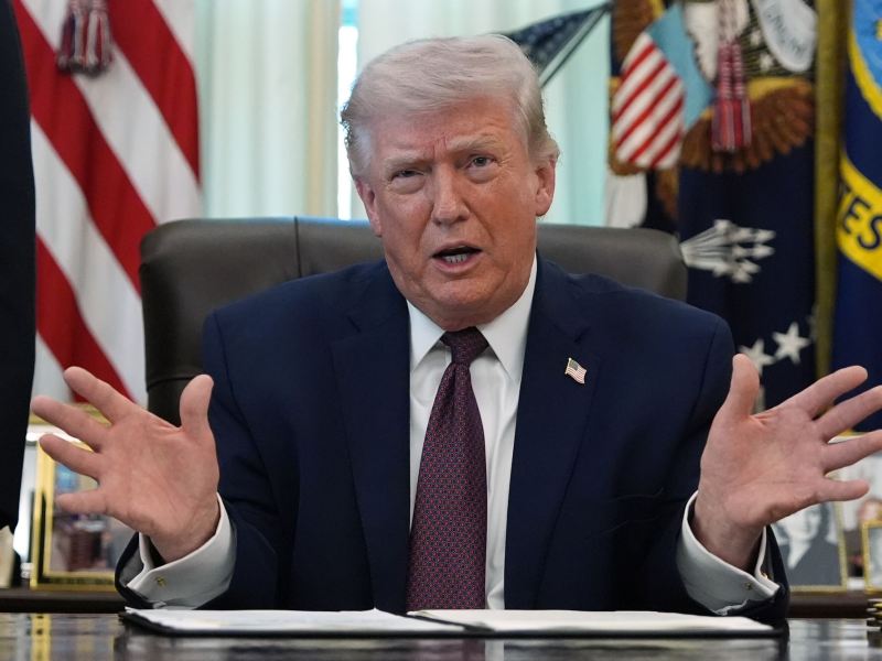 Donald Trump sitting at his desk, speaking with hands spread wide