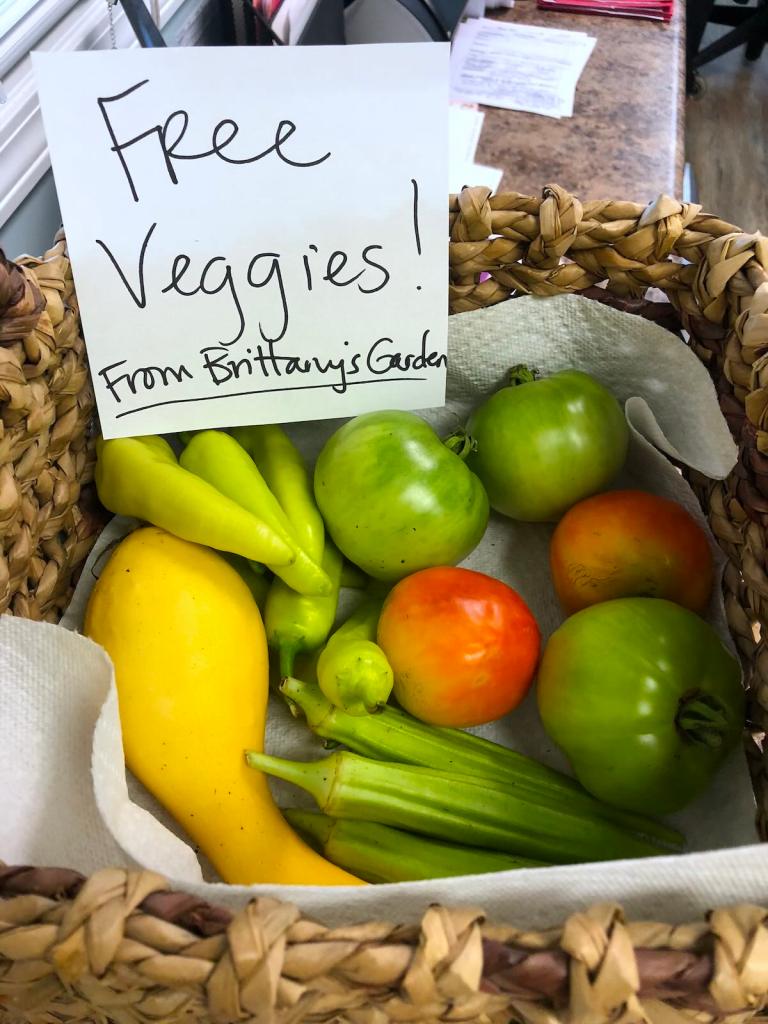 A basket of tomatoes, peppers, okra, and squash with a handwritten sign that says 'Free Veggies! From Brittany's Garden'