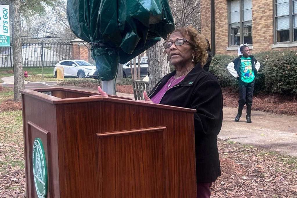 A woman in glasses speaking at a podium labeled 'Delta State University' located outside of a brick building