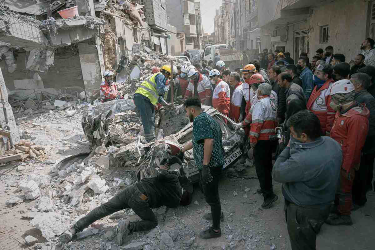 A group of people, some in protective gear, inspect a destroyed car in the middle of a street. Damaged buildings and rubble are in the background.