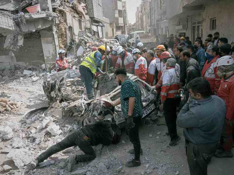 A group of people, some in protective gear, inspect a destroyed car in the middle of a street. Damaged buildings and rubble are in the background.