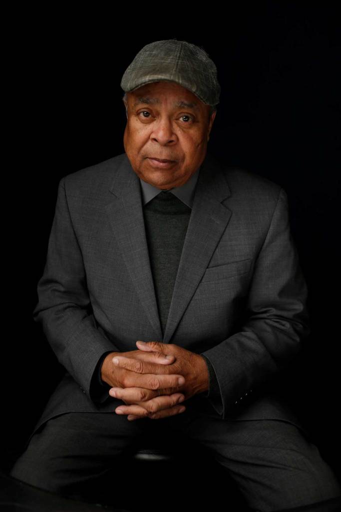 James Lap Baker wearing a grey suit and matching grey cap, makes a serious face while seated