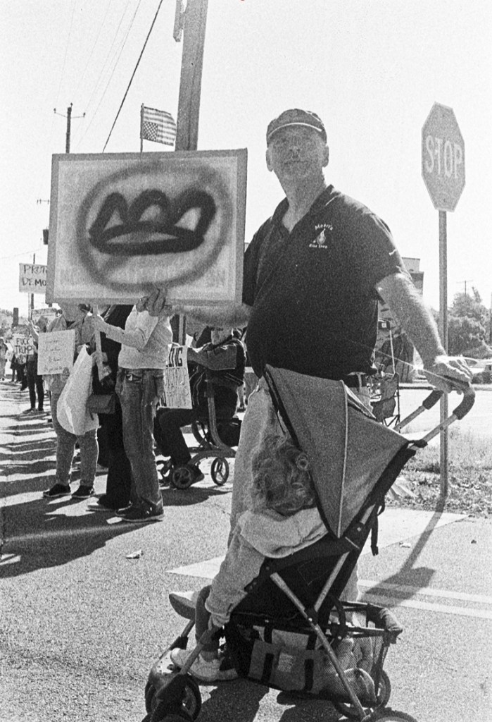 A man standing in a street with a child in a stroller holds up a sign with a crown and a round circle with a line through it over the crown