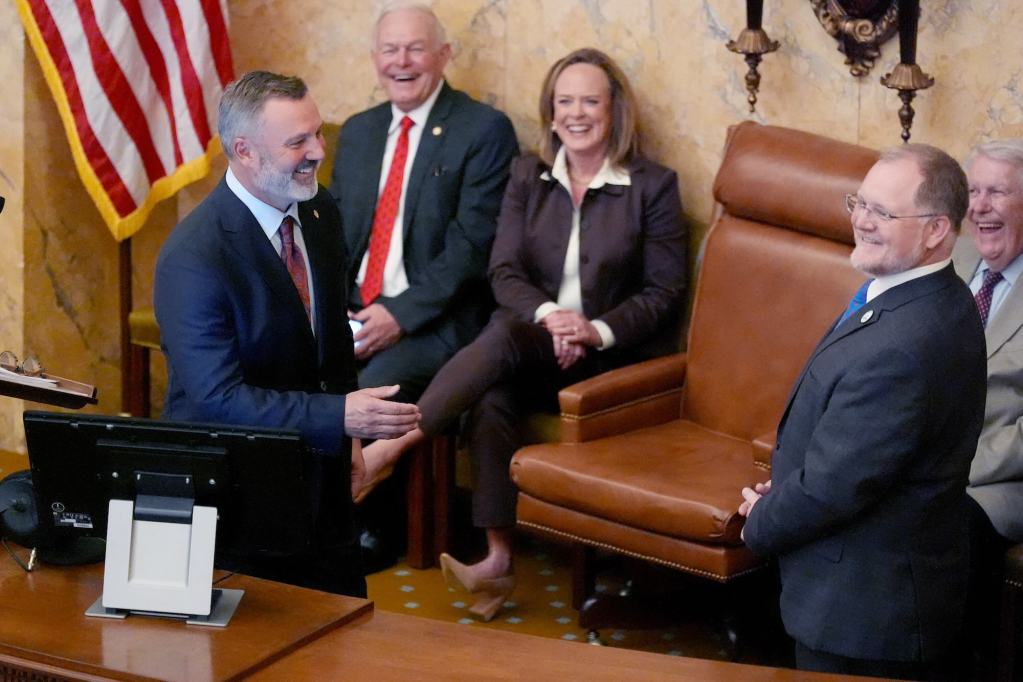 Legislatures, some sitting and some standing, speak and laugh