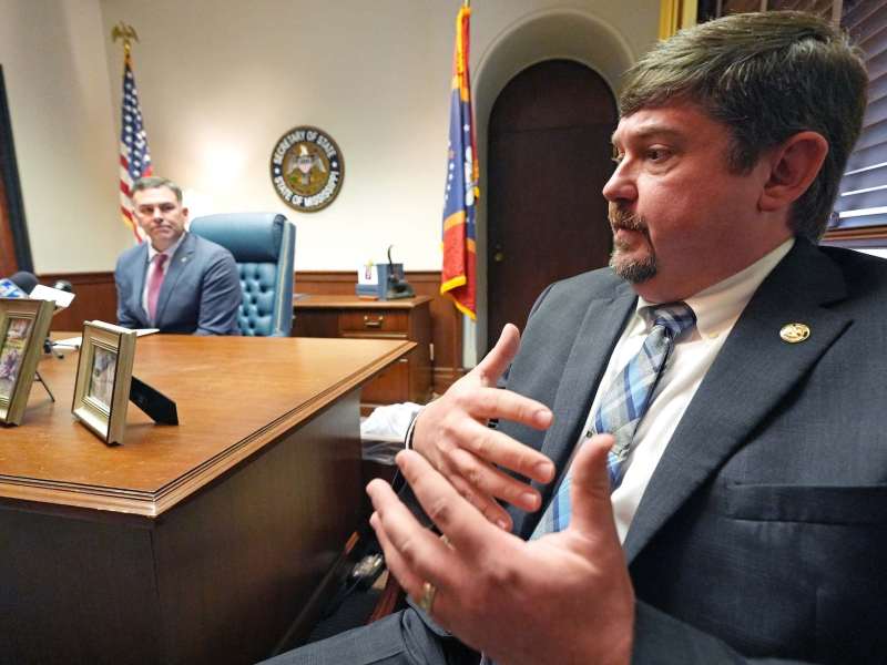 Jeremy England gestures with his hands in the foreground. Mississippi Secretary of State Michael Watson is sitting at a desk in the background.