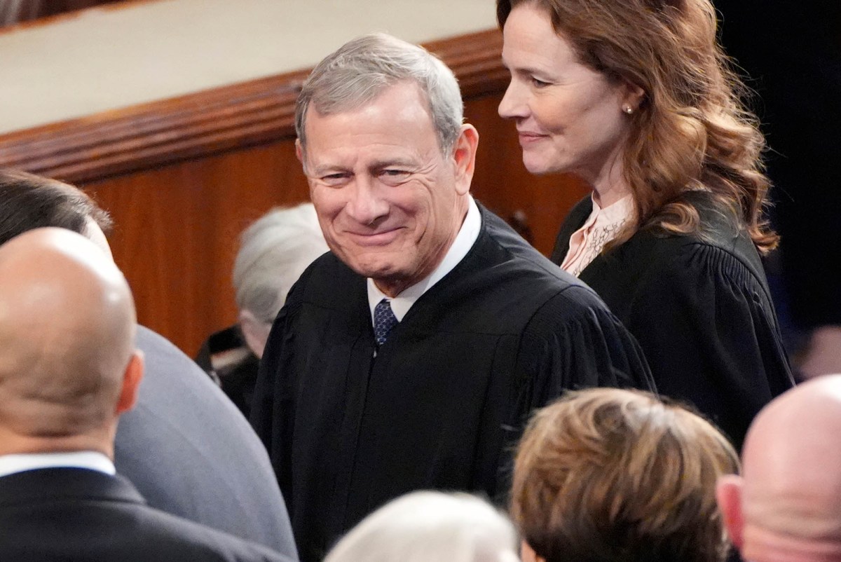 A closeup of Chief Justice of the Supreme Court John Roberts, in black robes, walking into a room of people